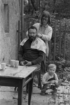 Schwarz-Weiß-Fotografie mit einer Frau, die einem Mann die Haare schneidet. Ein Kind sitzt daneben.