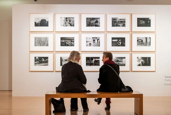 Zwei Besucherinnen sitzen auf einer Bank vor einer Wand mit Kunstwerken.