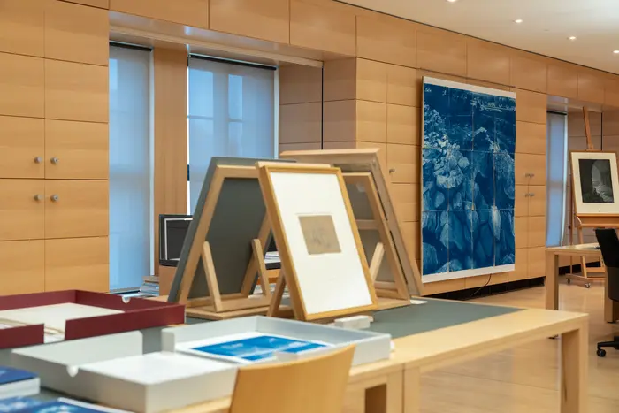 Ein Arbeitstisch mit aufgestellten gerahmten Bildern, im Hintergrund ein großes, blaues Gemälde an der Wand.