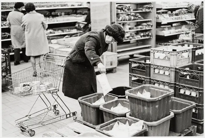 Schwarz-Weiß-Fotografie einer alten Frau mit Einkaufswagen im Supermarkt.