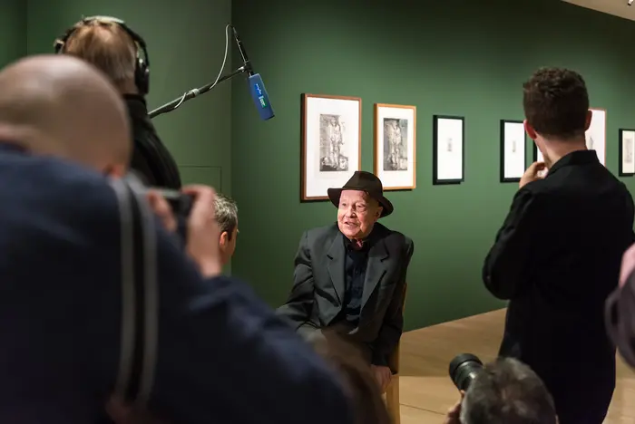 Ein Mann mit Hut sitzt in einem Ausstellungsraum und wird interviewt.