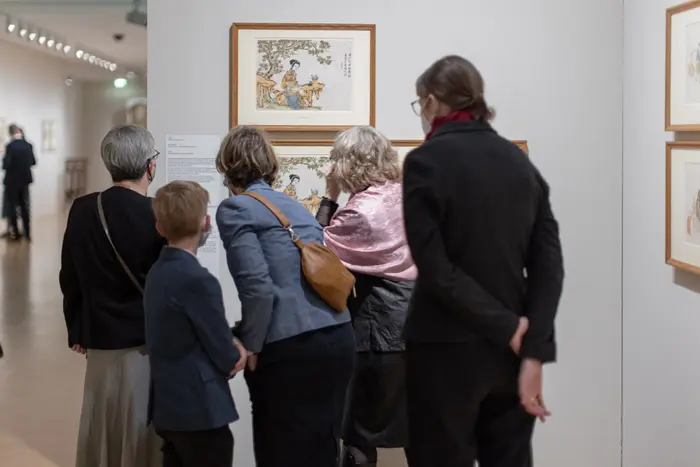 Mehrere Menschen betrachten gerahmte Zeichnungen in einem Ausstellungsraum.
