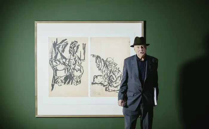 Ein Mann mit Hut steht vor einem gerahmten Kunstwerk an einer dunkelgrünen Wand.