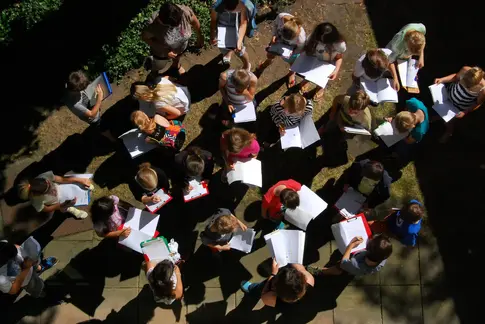 Ein Luftbild zeigt in der Natur sitzenden Menschen, die etwas auf ihren Mappen malen
