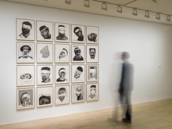 Eine Person betrachtet im Quadrat angeordnete Porträtzeichnungen an einer weißen Wand.