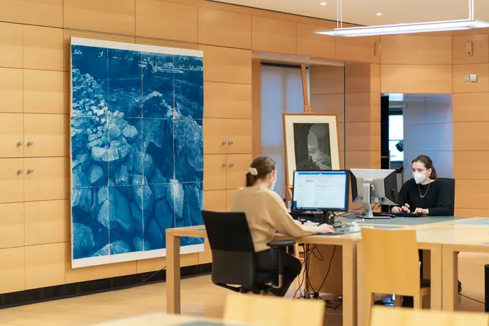 Zwei Personen Arbeiten an Computerbildschirmen mit einem großen blauen Kunstwerk an der Wand im Hintergrund.