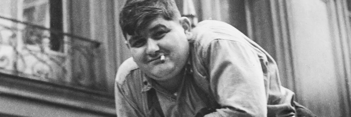 Schwarz-Weiß-Fotografie eines Mannes mit Zigarette im Mund, der auf einer großen Papierrolle lehnt.