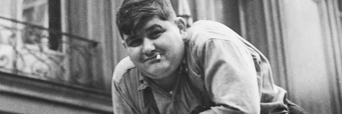 Schwarz-Weiß-Fotografie eines Mannes mit Zigarette im Mund, der auf einer großen Papierrolle lehnt.