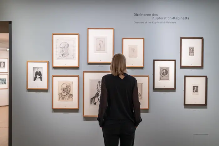 Eine Person betrachtet in Holz gerahmte Kunstwerke an einer grauen Wand.