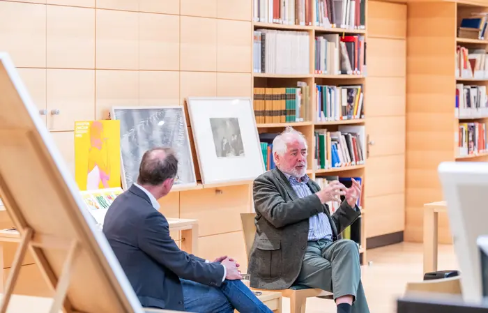 Zwei Männer diskutieren sitzend in einem Ausstellungsraum.