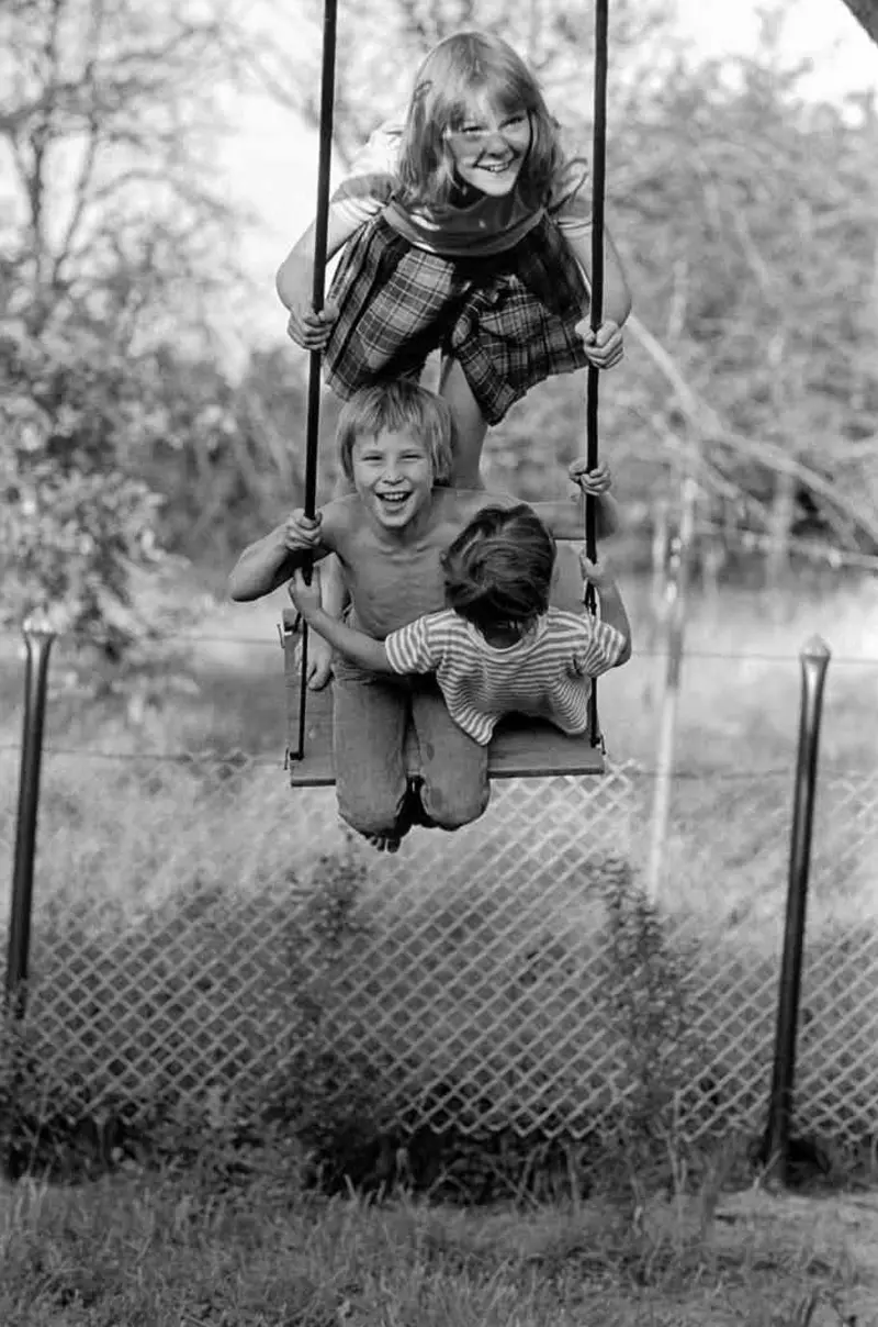 Schwarz-Weiß-Fotografie von drei lachenden Kindern auf einer Schaukel.