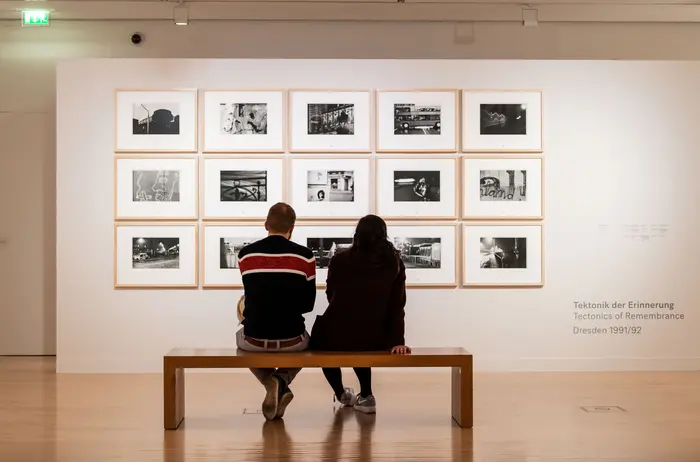 Zwei Besucher sitzen auf einer Bank vor einer Wand mit Kunstwerken.