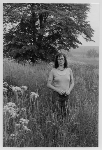 Schwarz-Weiß-Fotografie einer Frau, die in hohem Gras vor einem Baum steht.